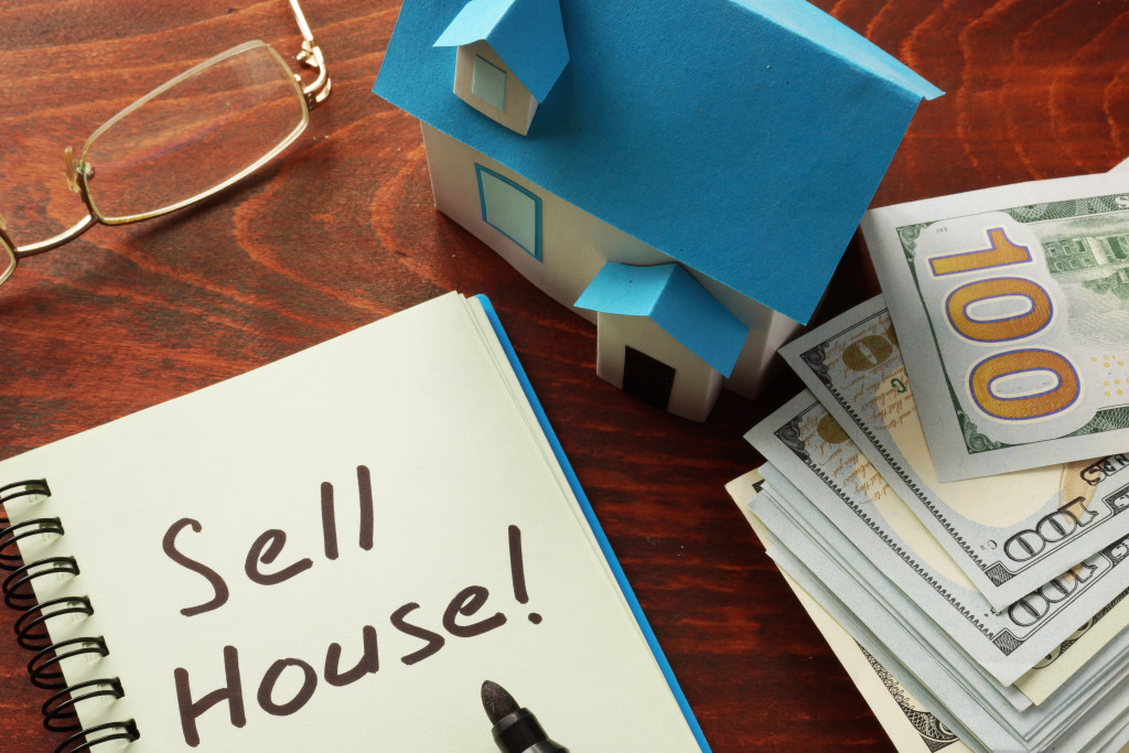 A notebook with the words Sell House! on table with a miniature house, money, and eyeglasses