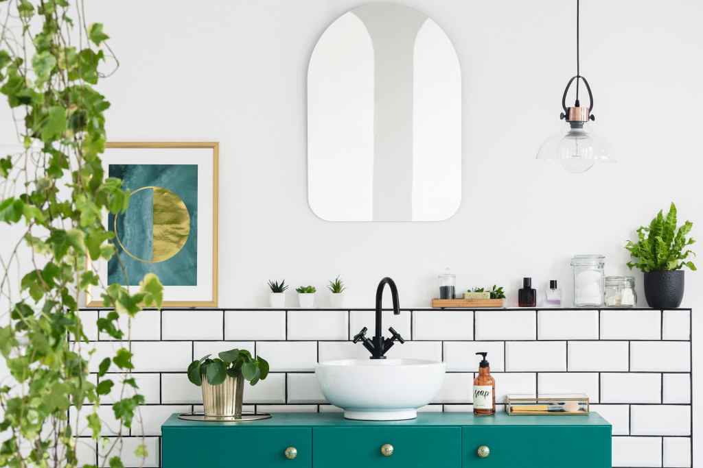 Mirror on white wall above green washbasin in bathroom interior with plants and poster